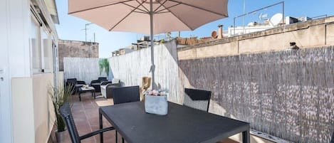En udendørs terrasse med et bord, stole, en stor parasol og en dekorativ plante.