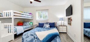 A bedroom with bunk beds, a flat-screen TV, a bedside table with a lamp, and a window with blinds.