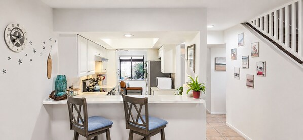 A modern kitchen with a central island, two chairs, a clock, and a staircase leading to another level.