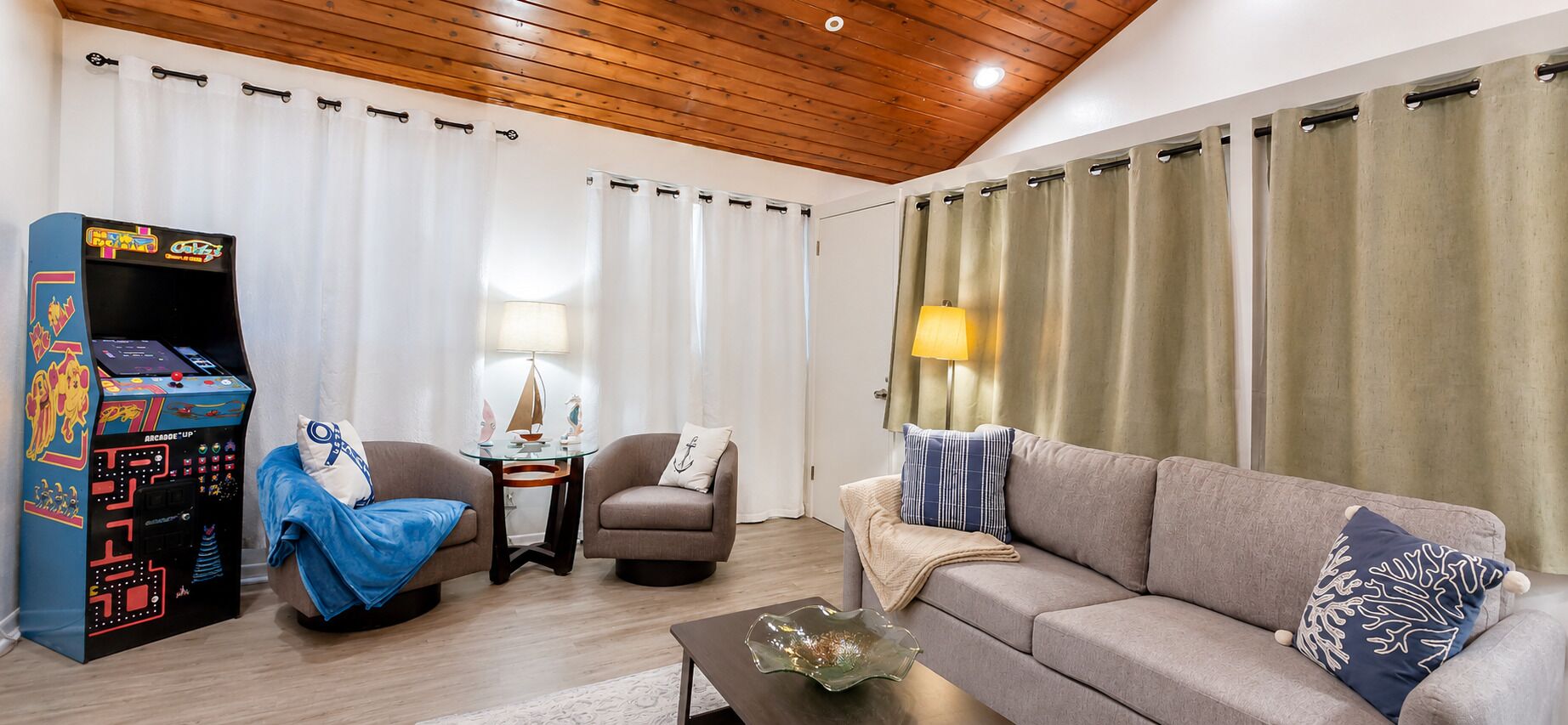 A living room with a sofa, armchairs, a coffee table, a lamp, curtains, and a retro arcade game.