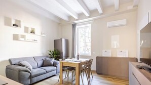 A modern living room with a grey sofa, wooden dining table, and a kitchen area with a fridge and cabinets. - Suite 53 Campofiore - with outdoor patio (Verona)