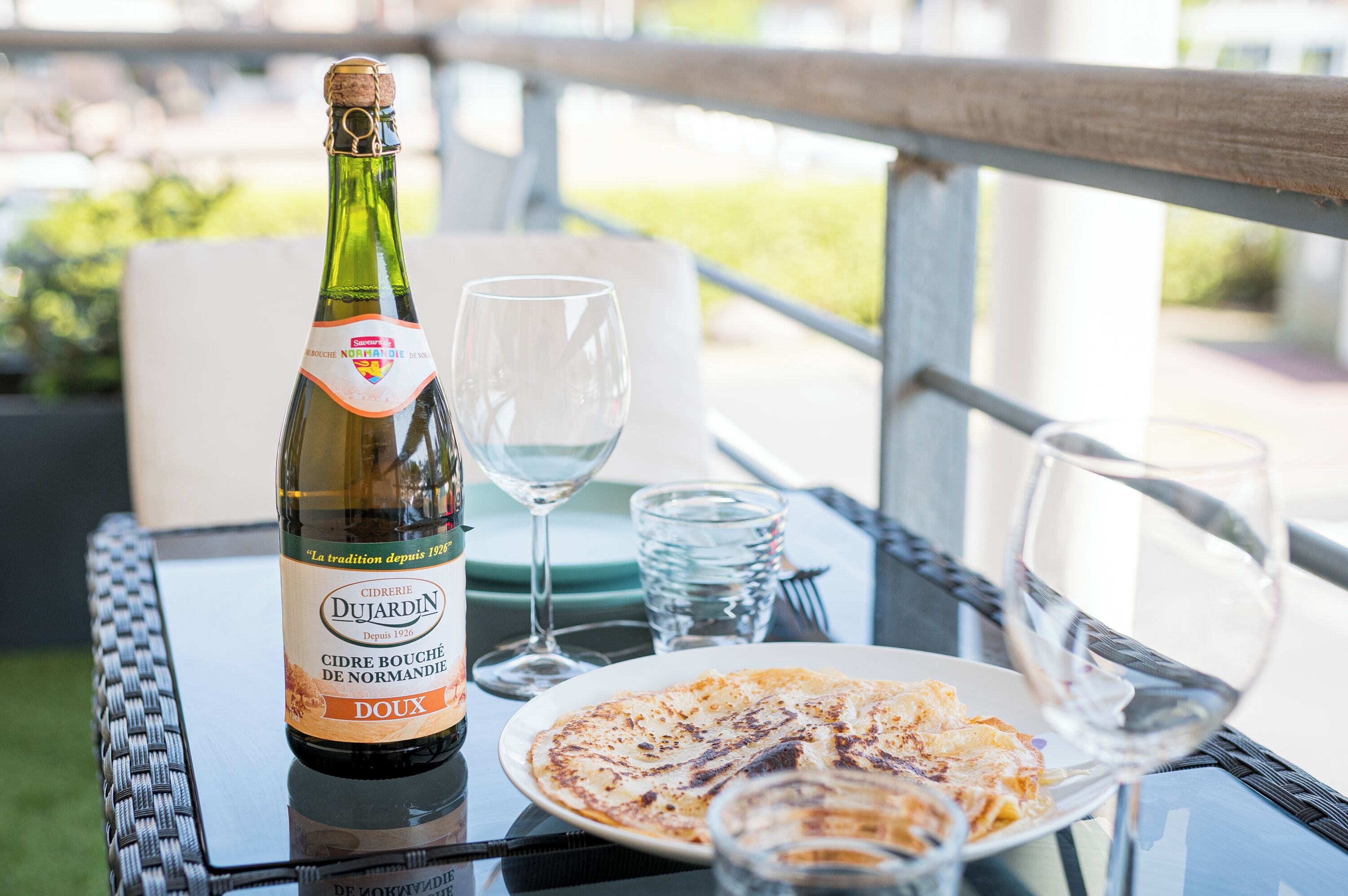 A bottle of Cidre Bouché de Normandie Doux wine, a glass, and a plate of food on an outdoor table.