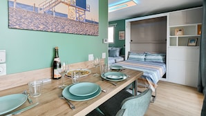 Un comedor con una mesa de madera dispuesta para dos, una cama con manta a rayas y una pared con un cuadro de playa.