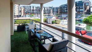 Un balcón con mesa lista para comer, con vistas a un puerto deportivo con barcos y edificios modernos.