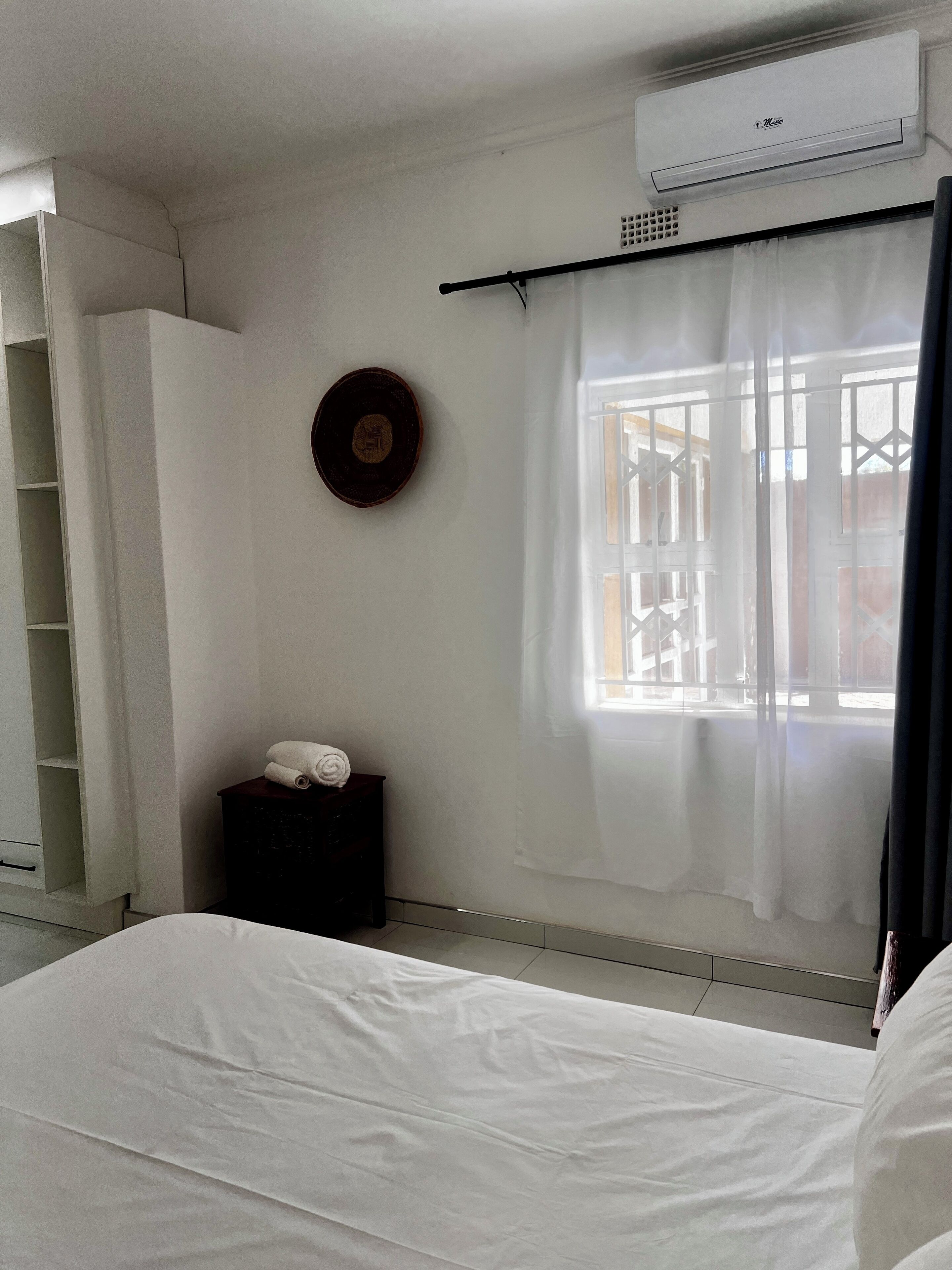 Habitación de hotel con cama, una mesita con toallas, aire acondicionado en la pared, ventana con cortina y un reloj de pared.
