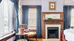 A room with a fireplace, a bed, a chair, a table, and a window with curtains.