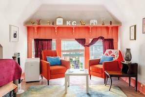 Une pièce avec deux fauteuils orange, une petite table blanche, un canapé rouge, un climatiseur blanc et une fenêtre avec des stores.