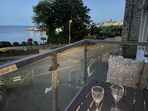 A balcony with a glass railing overlooking a street and a body of water. There are two empty wine glasses on the table. - Lifeboat Lookout (Tenby)