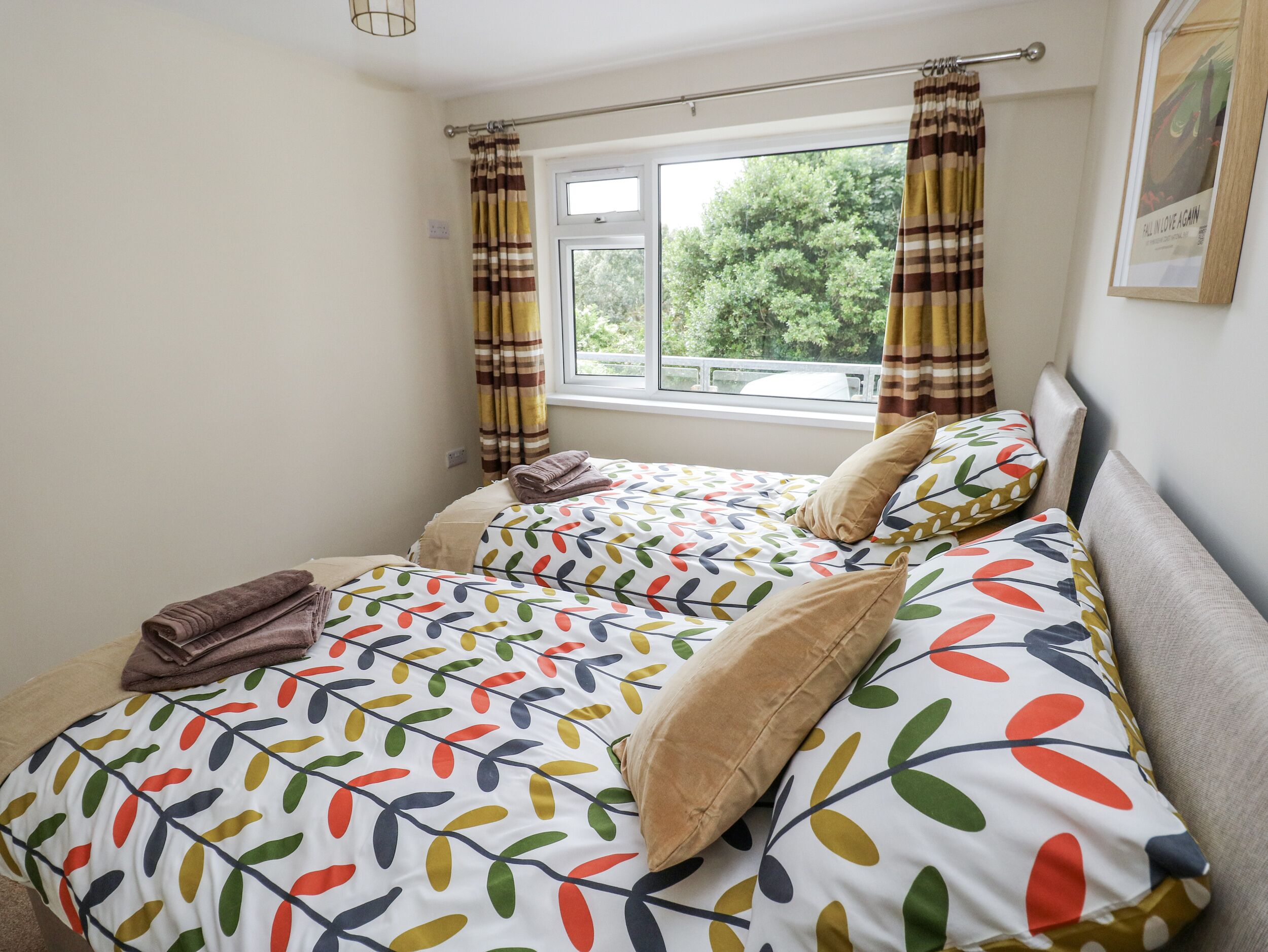 A bedroom with two beds, striped curtains, a window with a view of trees, and a framed picture on the wall.
