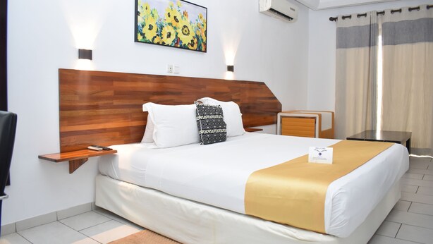 Hotelkamer met een houten hoofdbord, een bed met wit beddengoed en een gouden plaid, een klein nachtkastje, een nachtlampje, een lijst met bloemkunst en een bordje met de tekst 'Welkom'.