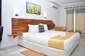 Une chambre d’hôtel avec une tête de lit en bois, un lit avec une literie blanche et un plaid doré, une petite table de chevet, une commode, une œuvre d’art florale encadrée et une pancarte « Bienvenue ».