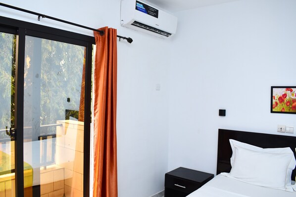 Une chambre à coucher comprenant un lit, une table de chevet, un climatiseur fixé au mur, une porte-fenêtre coulissante et un tableau accroché au mur.