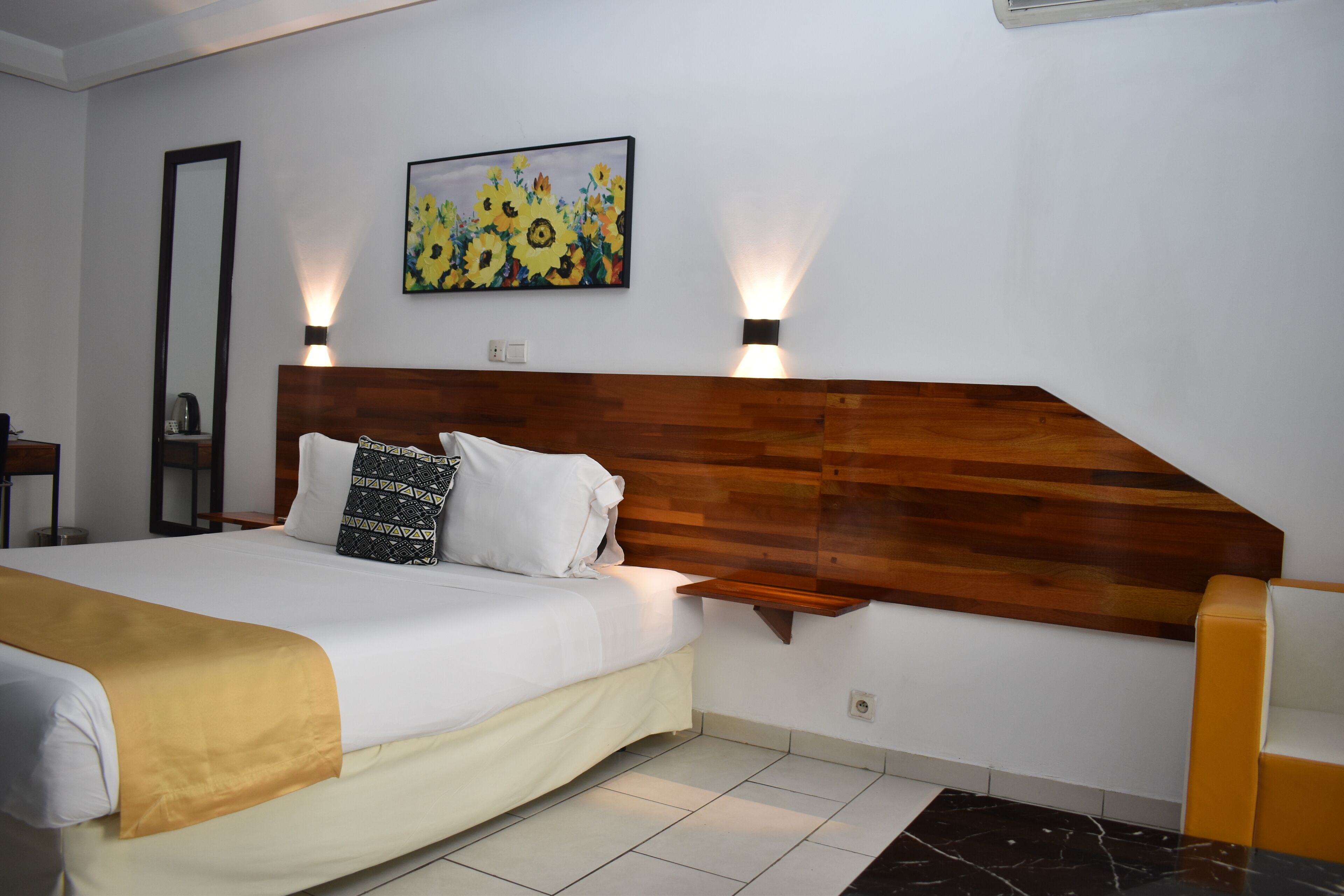 A neatly made bed with a wooden headboard, a painting of sunflowers, and two wall-mounted lights.