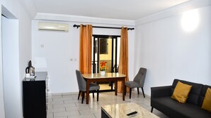 Een moderne woonkamer met een zwarte bank, een salontafel, een eettafel met twee stoelen, en een klein keukenhoekje met een zwart kastje en een waterkoker.