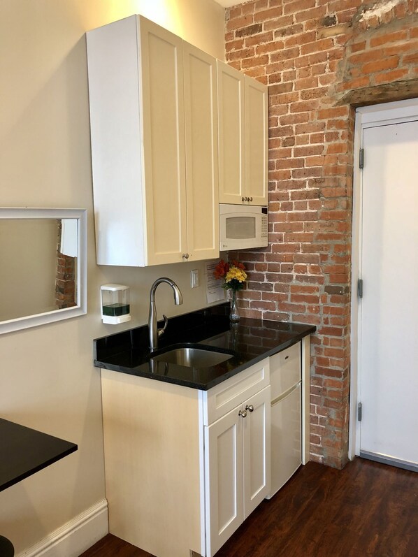 A compact kitchen with white cabinets, a black countertop, a stainless steel sink, and a microwave. - The Grand NYC (New York)