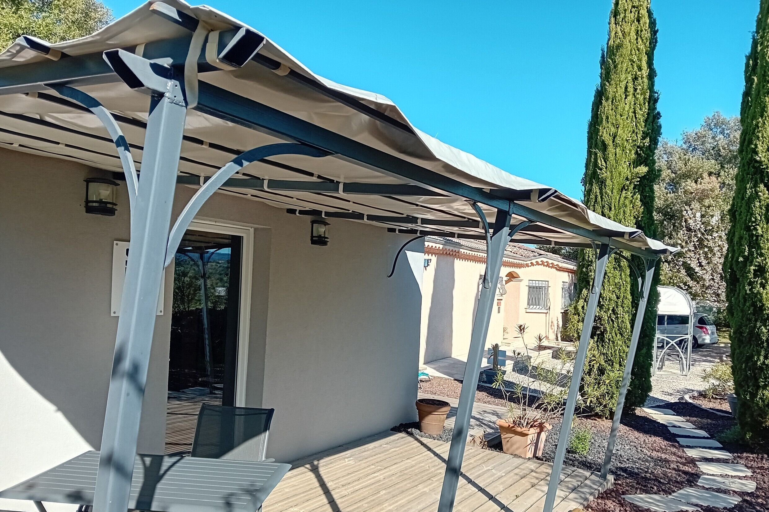 Ett takförsett utomhusområde med en pergola, ett bord och stolar, en gångväg som leder till en byggnad och höga cypressträd.