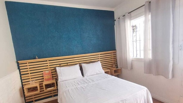 Een slaapkamer met een bed, houten hoofdbord, een klein nachtkastje, een nachtlampje, een raam met gordijnen en een plafondlamp.