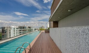 A rooftop swimming pool with a wooden deck, a concrete wall, and a view of surrounding buildings. - Your Urban Retreat Near the Subway - Iby1613 (São Paulo)