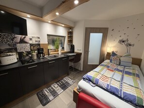 Une chambre d’hôtel de taille réduite, équipée d’une kitchenette, d’un lit, d’un bureau et d’une porte.