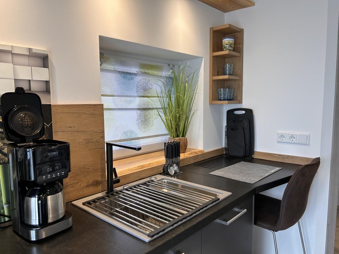 A modern kitchen with a built-in coffee maker, a window with a plant, and a wooden shelf with decorative items.