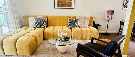 A living room with a yellow sectional sofa, a glass coffee table, and a dark armchair.