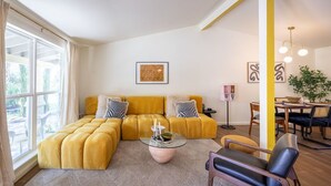 Una sala de estar con un sofá amarillo, una mesa de centro de vidrio y una silla negra. Hay una zona de comedor con una mesa y sillas.
