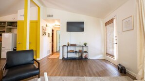 Una sala de estar con un sofá de cuero negro, un televisor de pantalla plana montado en la pared, piso de madera y una mesita con objetos decorativos.