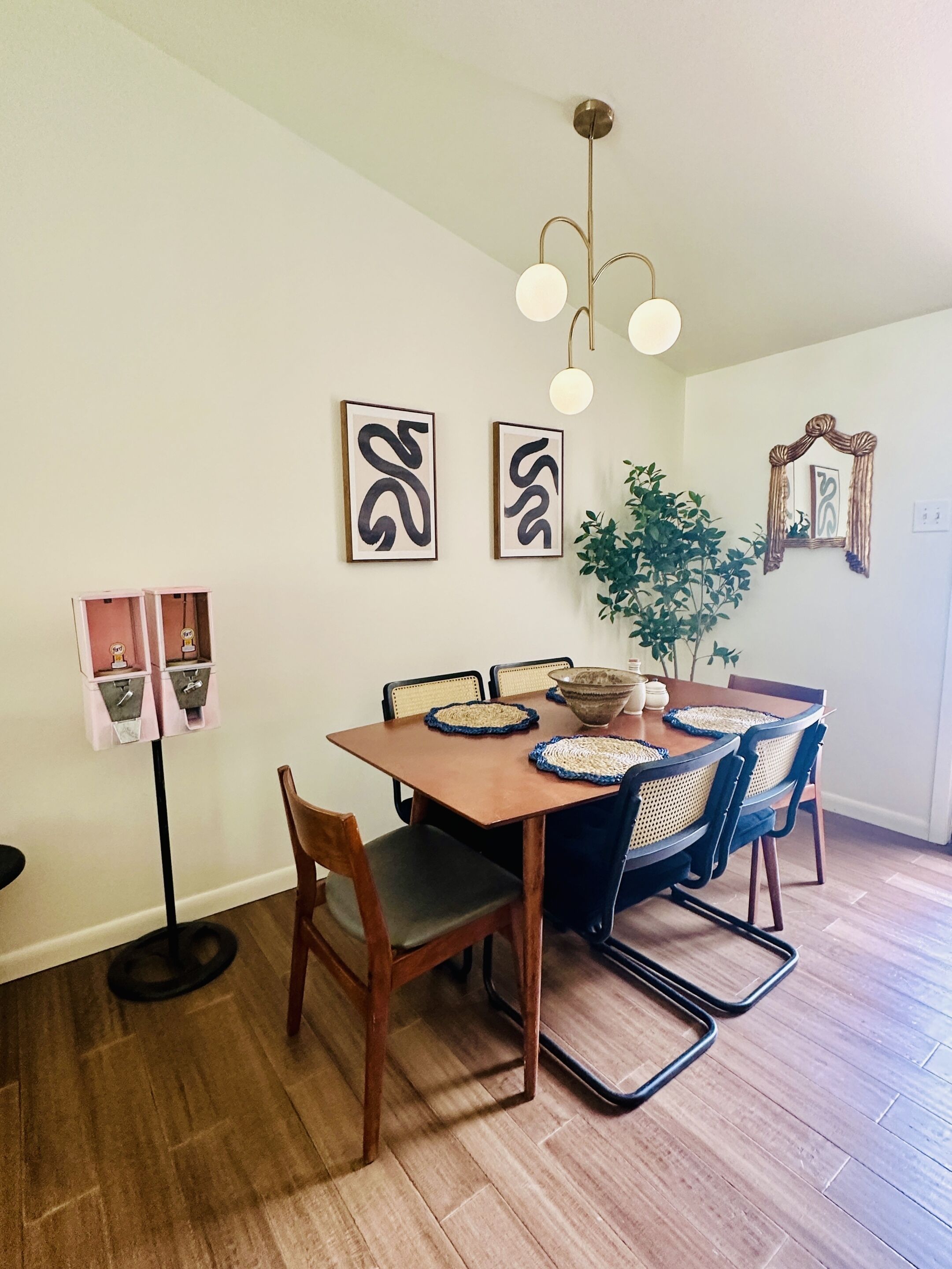 A dining area with a wooden table, four chairs, two wall-mounted abstract art pieces, a hanging light fixture, and a potted plant.