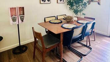 A dining area with a wooden table, four chairs, two wall-mounted abstract art pieces, a hanging light fixture, and a potted plant.