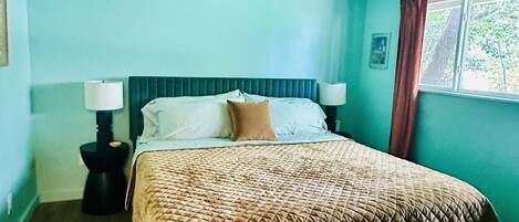 Un dormitorio con una cama, dos lámparas de noche, un tapete estampado, un manto blanco mullido, un candelabro en el techo y una ventana con cortinas.