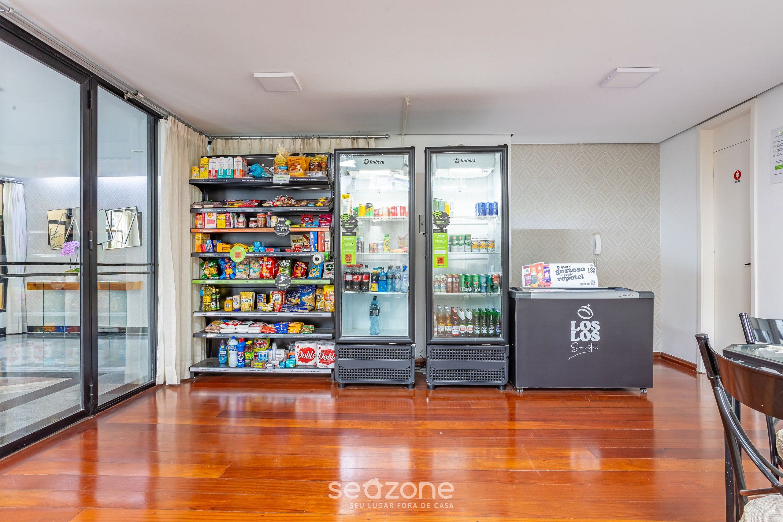 Een hotel lobby met een glazen deur die uitkomt op een balkon, een automaatgedeelte met snacks en drankjes, en een receptiebalie.