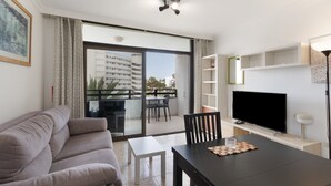 A living room with a sofa, a dining table, a TV, and a balcony with a view of buildings.