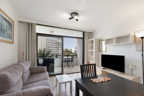 A living room with a sofa, a dining table, a TV, and a balcony with a view of buildings.