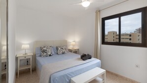 A bedroom with a bed, bedside tables, a ceiling fan, a window, and a view of buildings.