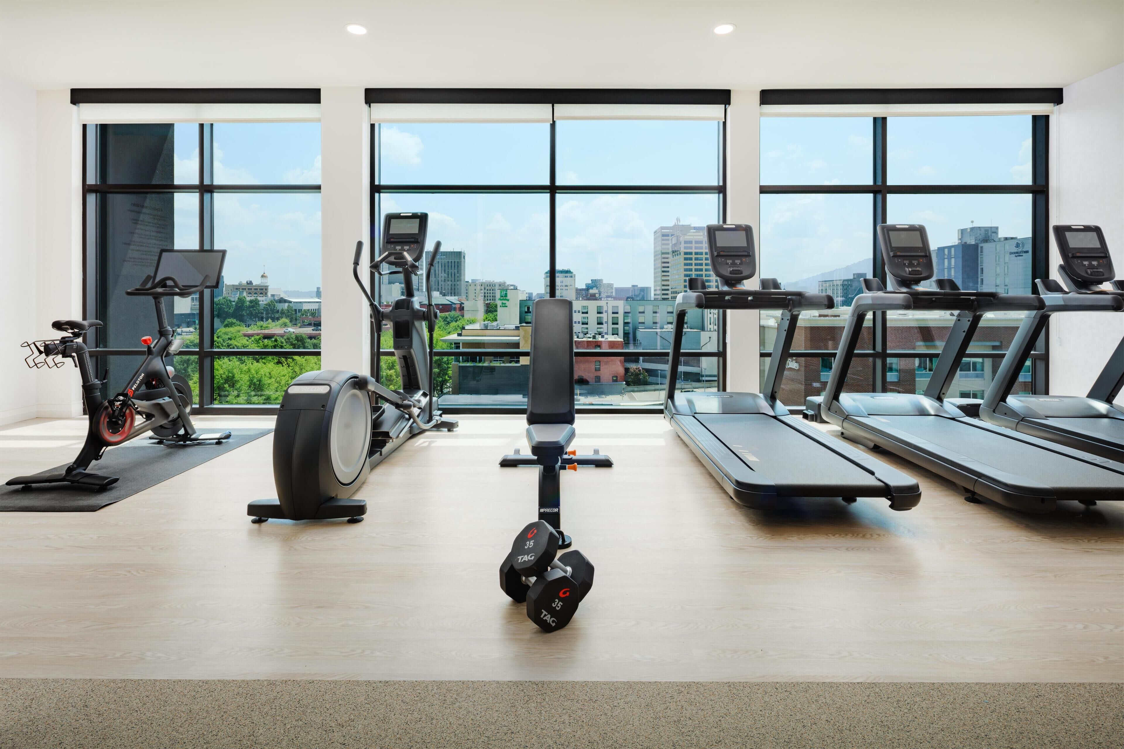 A gym with cardio machines, dumbbells, and a view of the city through large windows.