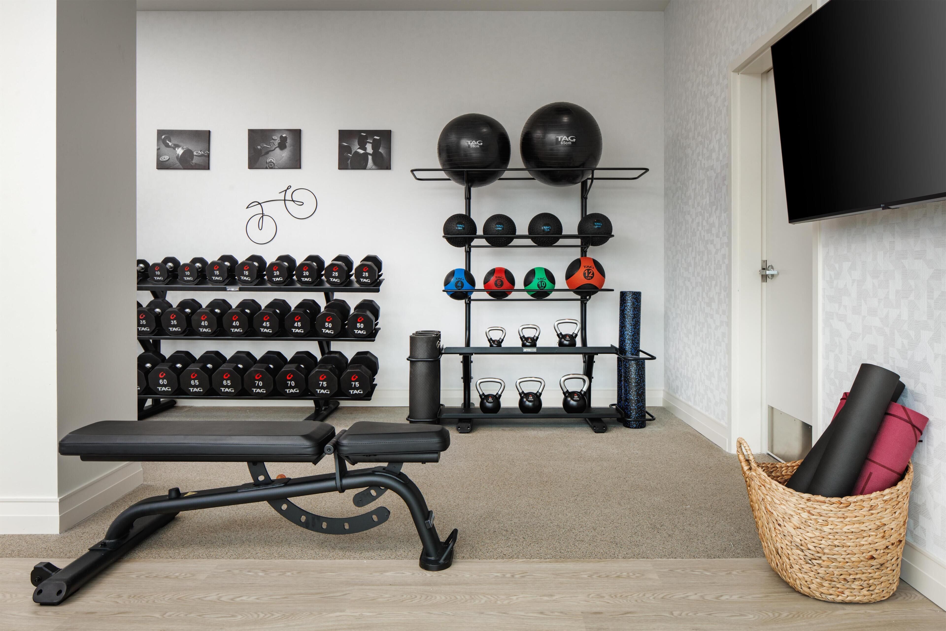 Un gimnasio en casa con una banca, mancuernas, kettlebells y un televisor montado en la pared.
