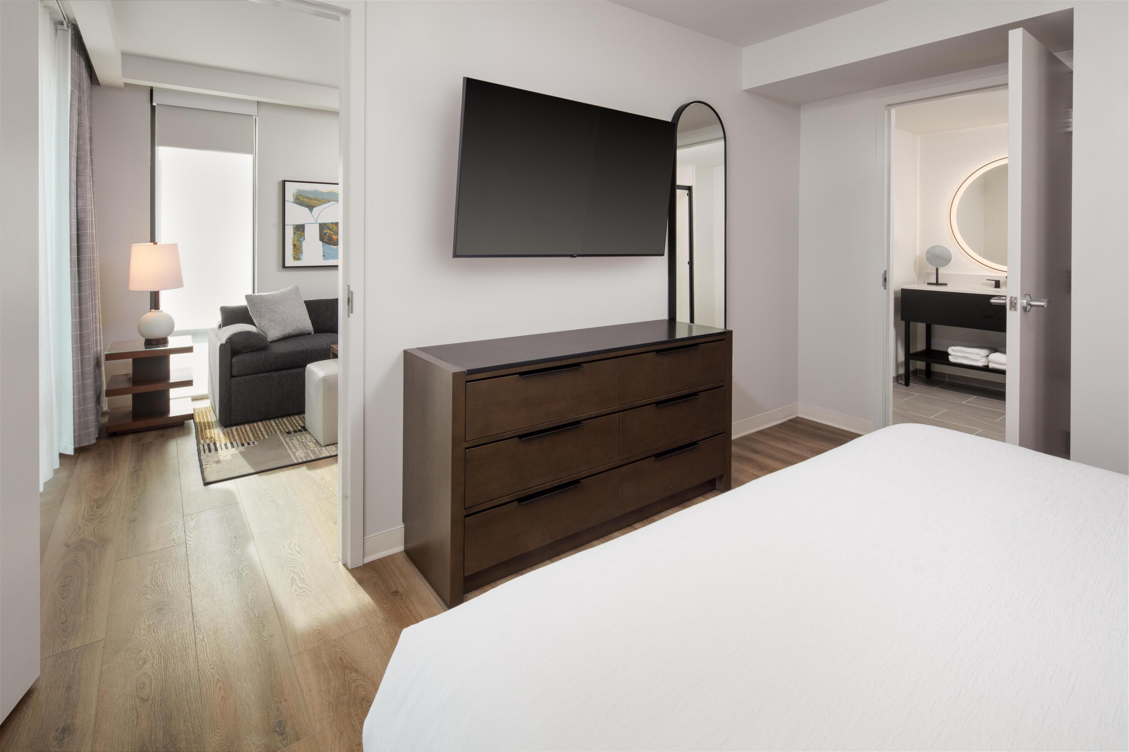 A modern bedroom with a large bed, a dresser, a flat-screen TV, and a vanity with a mirror.