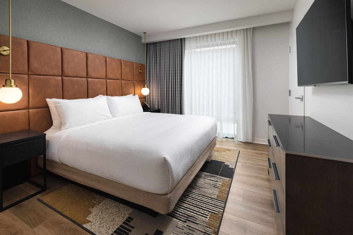 Una habitación de hotel moderna con una cama grande, un televisor de pantalla plana, un cabecero de madera y un amplio ventanal con cortinas.