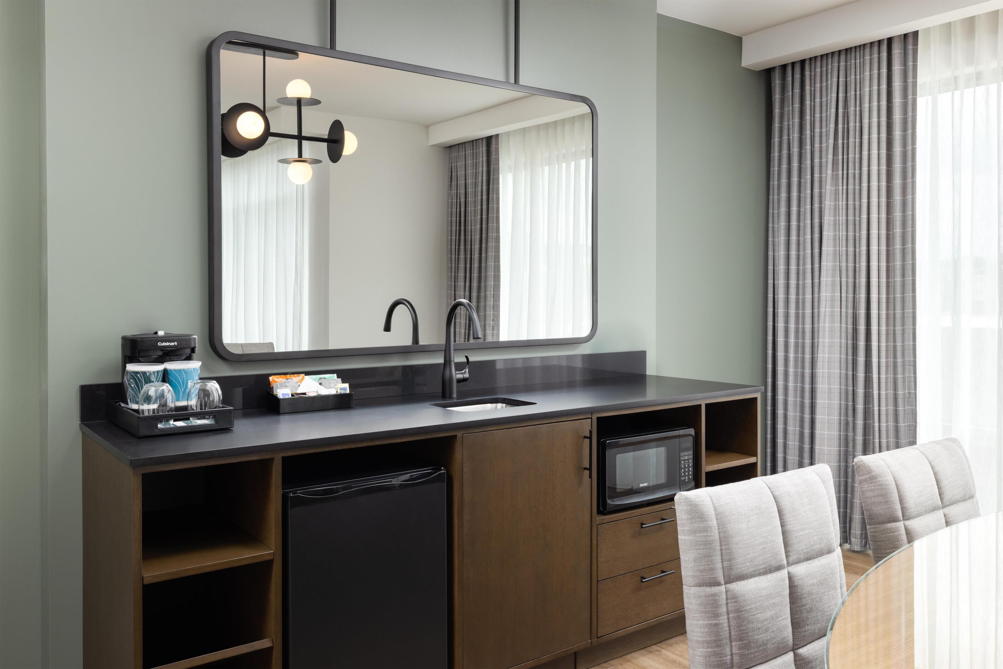 Une salle de bain moderne avec un grand miroir, un lavabo, une machine à café et un four à micro-ondes. On y trouve deux chaises et une fenêtre avec des rideaux.