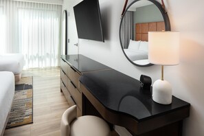 Una habitación de hotel con cama, escritorio, silla, televisor, lámpara y espejo.