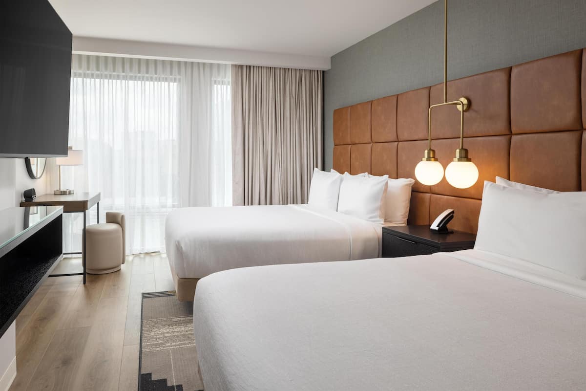 Habitación de hotel con dos camas, un escritorio, un televisor y un ventanal grande con cortinas.