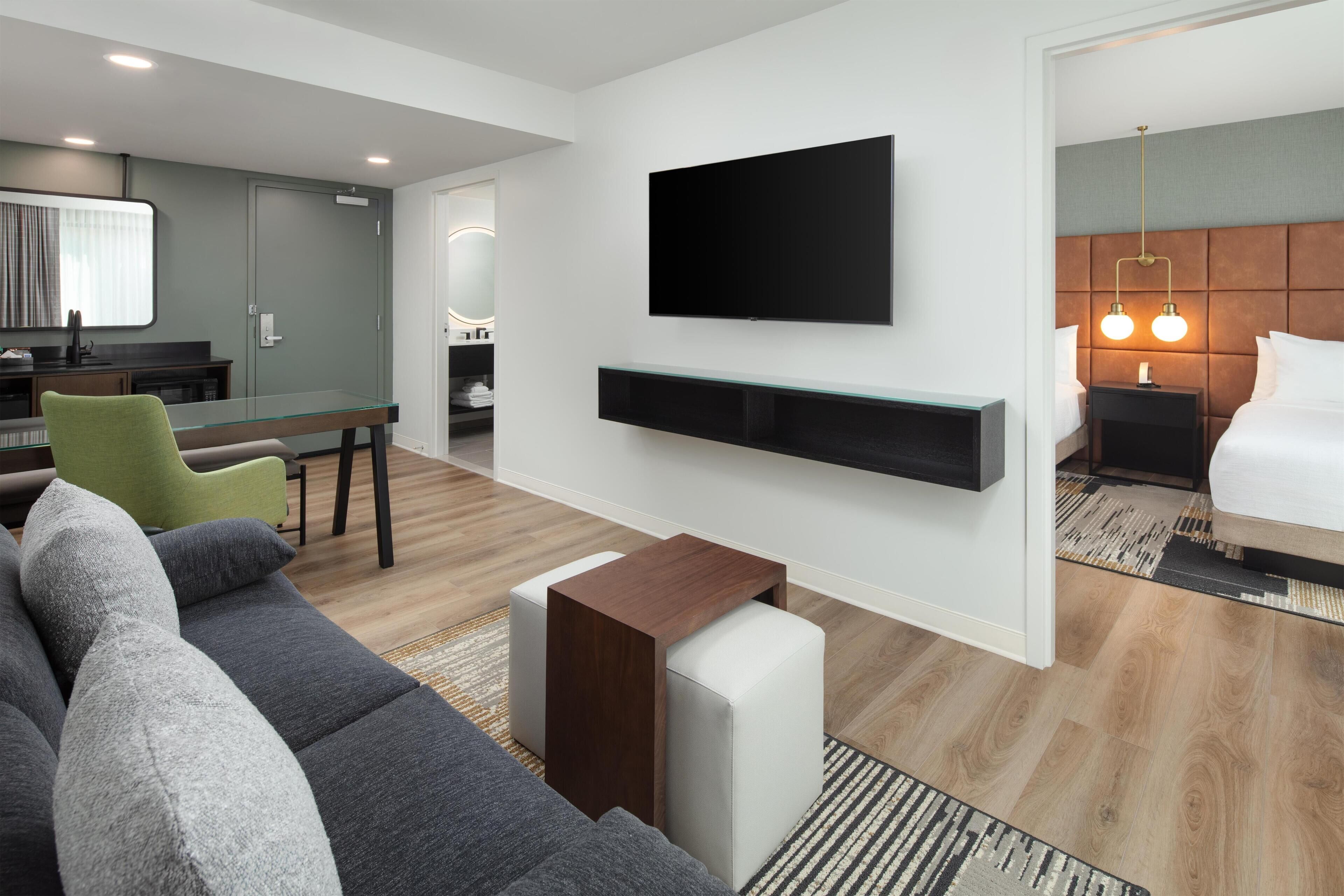 A modern hotel room with a flat-screen TV mounted on the wall, a sofa, a small wooden side table, and a bed with a headboard.
