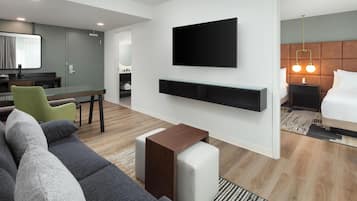 A modern hotel room with a flat-screen TV mounted on the wall, a sofa, a small wooden side table, and a bed with a headboard.