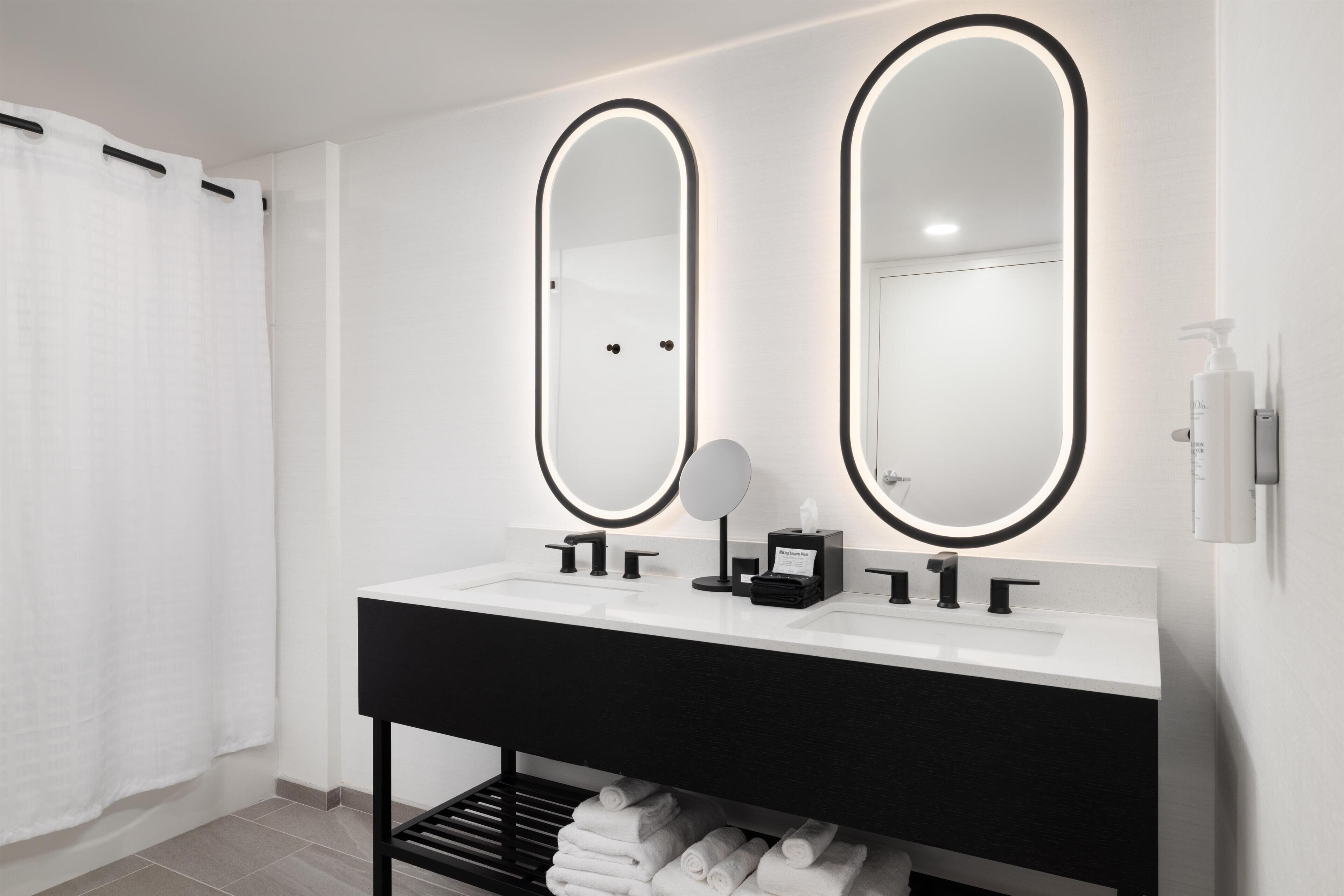 A modern bathroom with a black vanity, two large mirrors with illuminated frames, a white sink, and a towel rack with neatly folded towels.