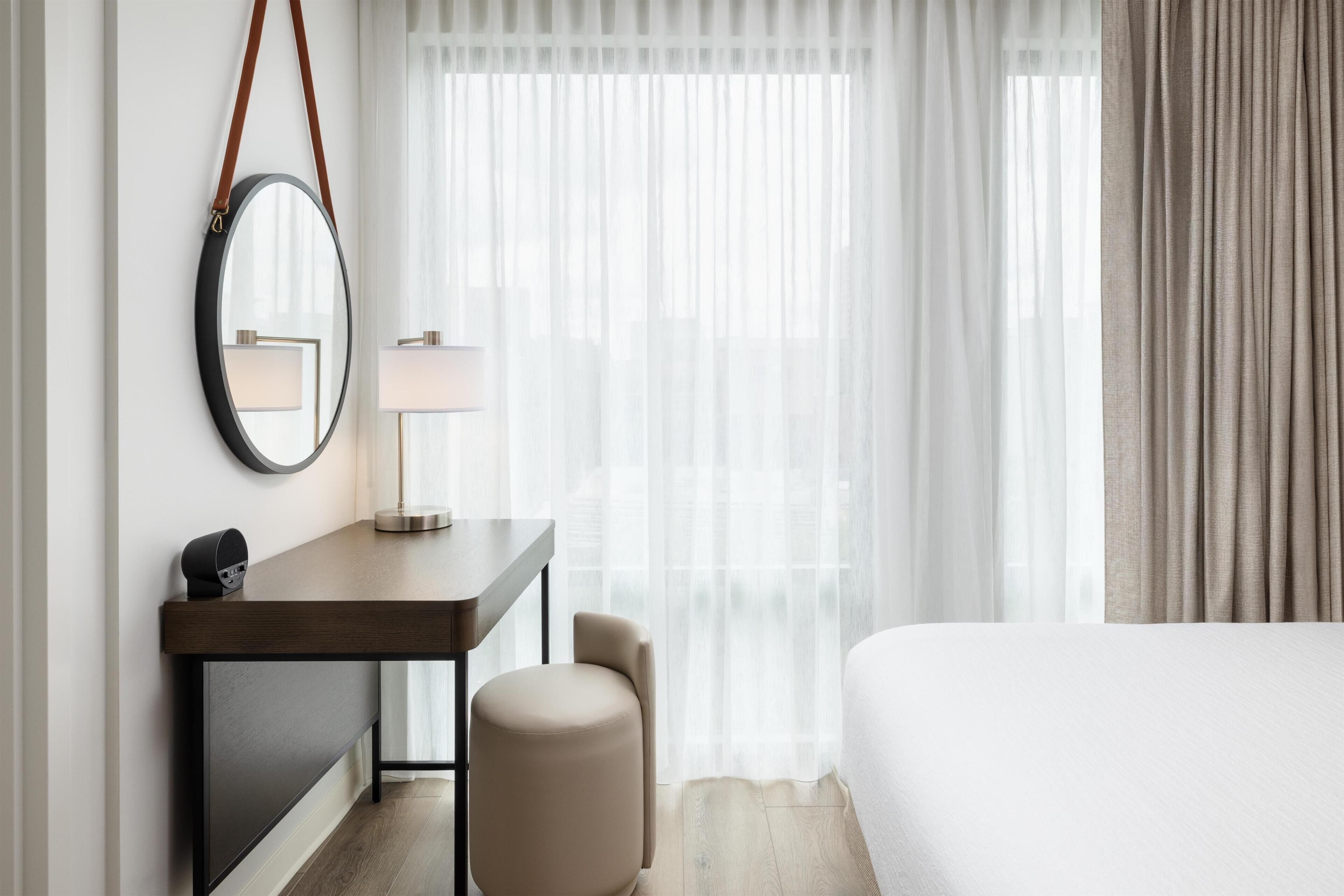 A hotel room with a bed, a desk with a lamp, a round mirror, and a chair.