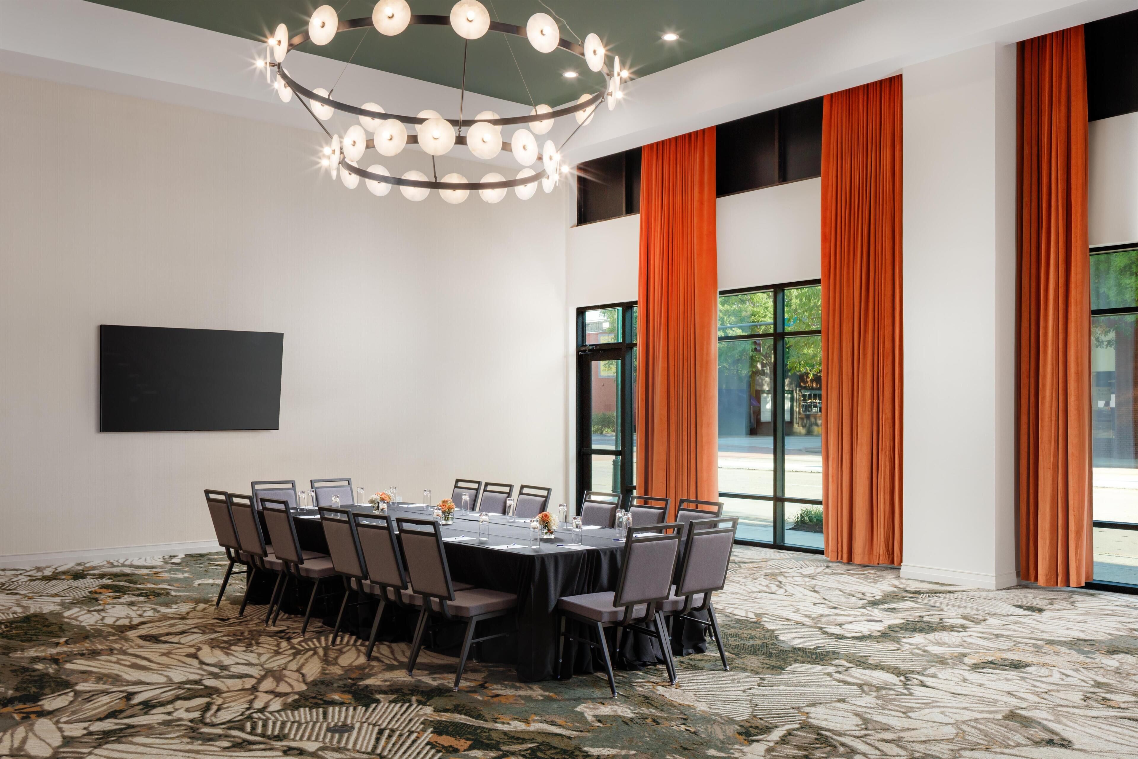 A conference room with a round table, chairs, a chandelier, and large windows with orange curtains.