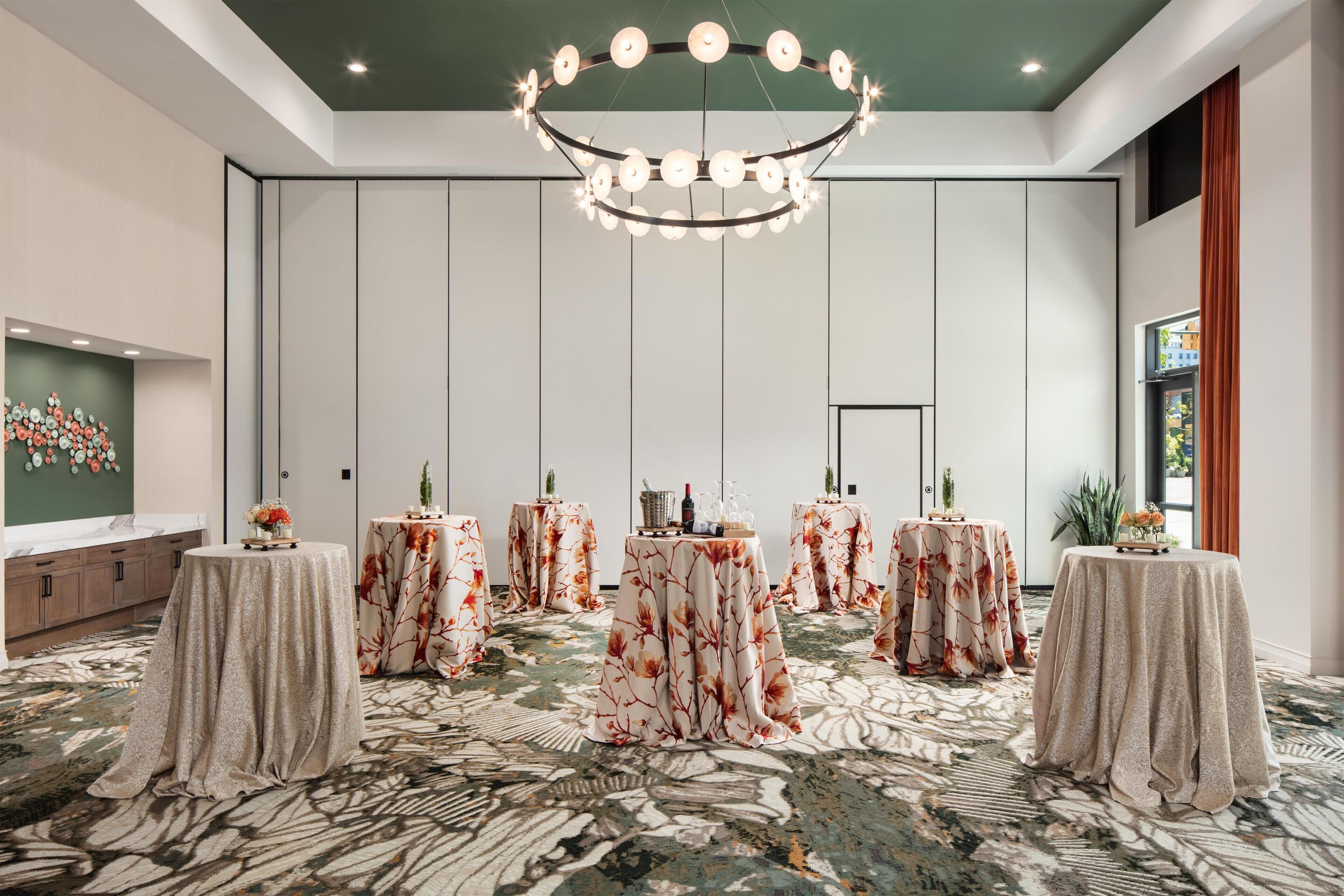 A hotel room with a large chandelier, multiple round tables with floral tablecloths, a large mirror, and a patterned carpet.