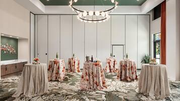 A hotel room with a large chandelier, multiple round tables with floral tablecloths, a large mirror, and a patterned carpet.