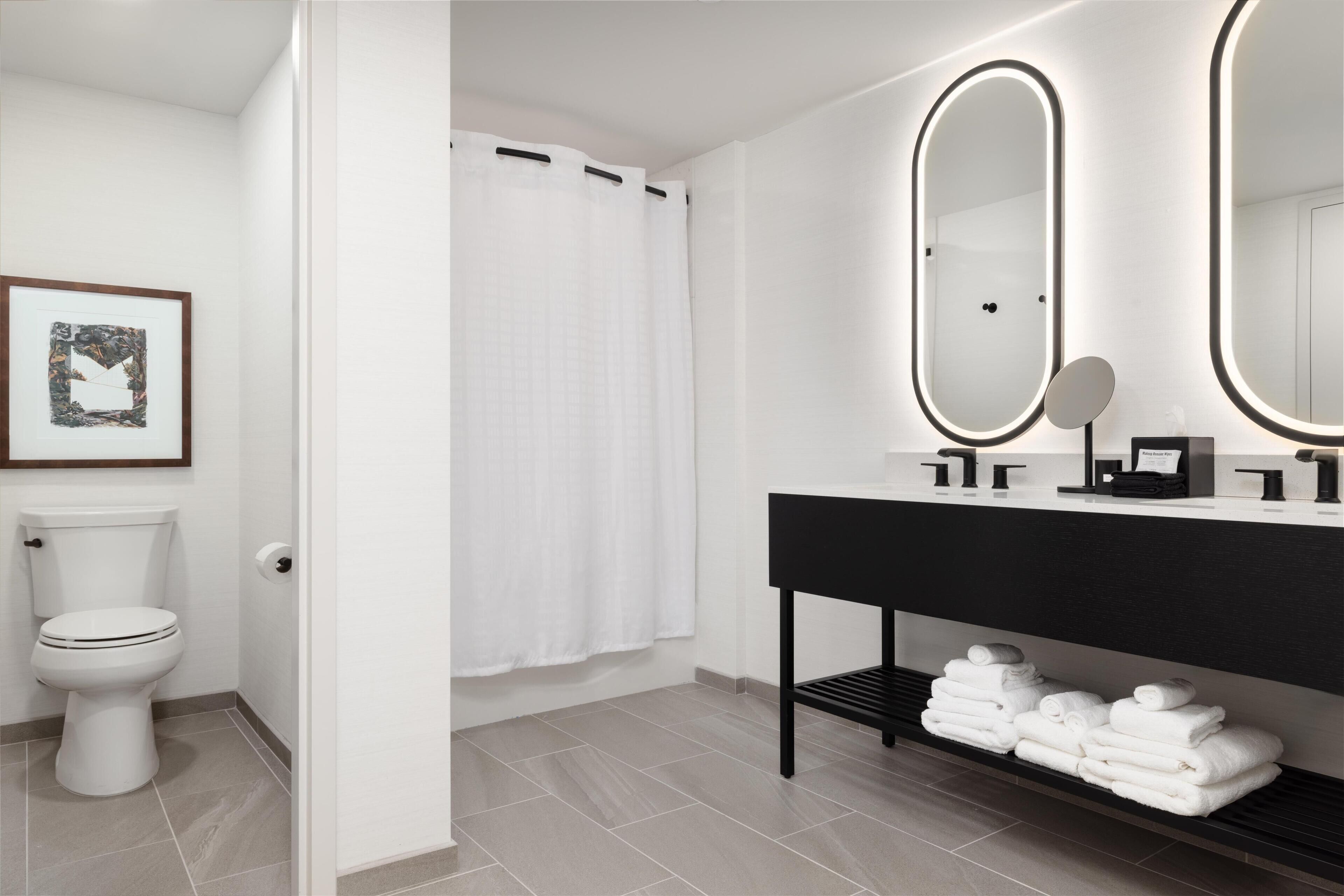 Une salle de bain moderne avec des toilettes, un rideau de douche, un meuble-lavabo avec un miroir et des serviettes soigneusement pliées sur une étagère.