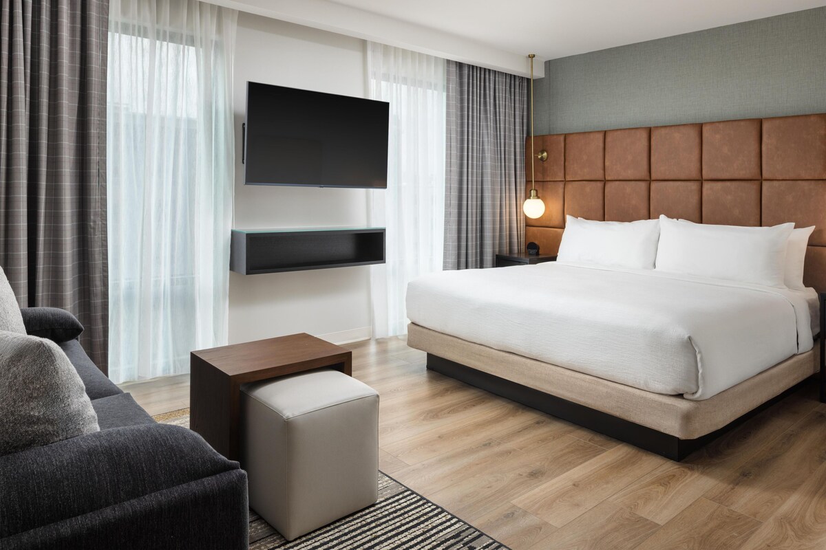Une chambre d’hôtel moderne dotée d’un grand lit, d’un téléviseur à écran plat fixé au mur, d’une table de chevet en bois et d’un coin salon avec un canapé et un pouf.